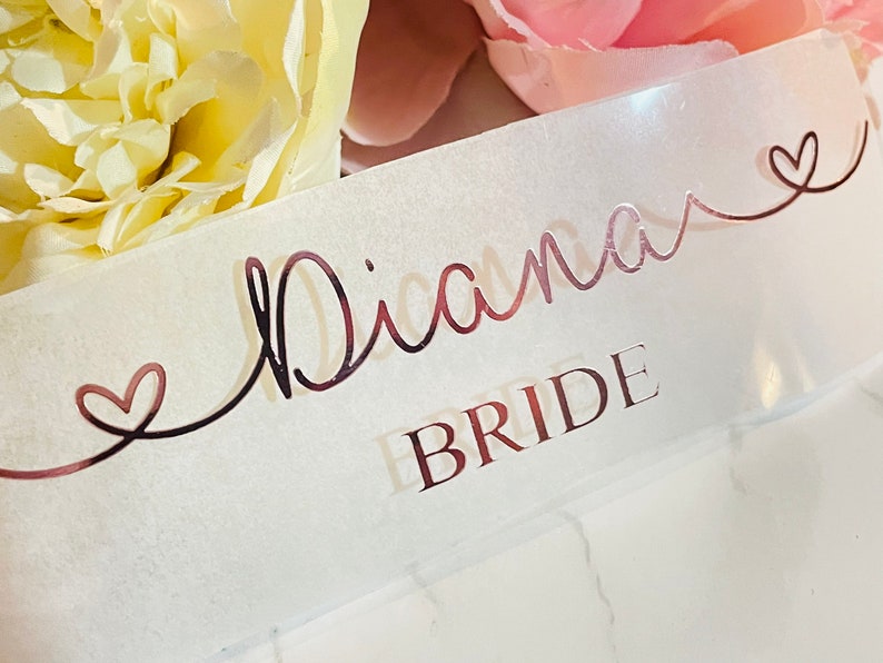 May include: A white satin ribbon with the name "Diana" written in rose gold script with a heart on either side. The words "Bride" are written in rose gold below the name.