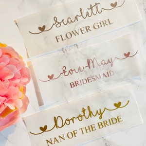 May include: Three white labels with gold and rose gold lettering on a marble background. The labels read "Scarlett Flower Girl", "Love-May Bridesmaid", and "Dorothy Nan of the Bride".