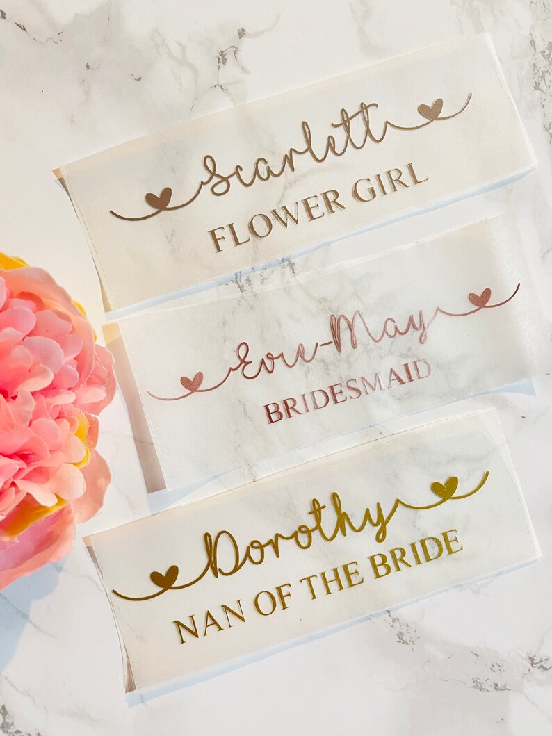 May include: Three white labels with gold and rose gold lettering on a marble background. The labels read "Scarlett Flower Girl", "Love-May Bridesmaid", and "Dorothy Nan of the Bride".