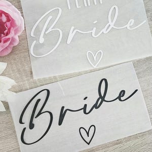 K&ouml;nnte beinhalten: Zwei wei&szlig;e Papierb&ouml;gen mit den Worten "TEAM BRIDE" in Schreibschrift. Einer hat wei&szlig;en Text und ein Herz, der andere hat schwarzen Text und ein Herz. Eine rosa Blume ist oben links zu sehen.