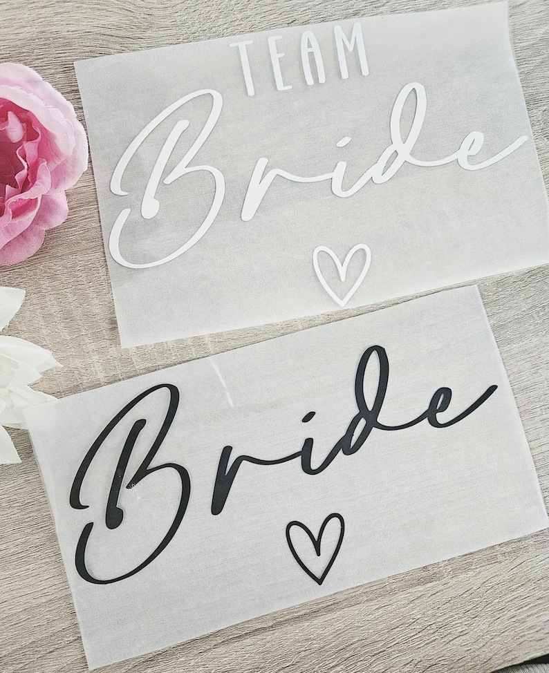 K&ouml;nnte beinhalten: Zwei wei&szlig;e Papierb&ouml;gen mit den Worten "TEAM BRIDE" in Schreibschrift. Einer hat wei&szlig;en Text und ein Herz, der andere hat schwarzen Text und ein Herz. Eine rosa Blume ist oben links zu sehen.