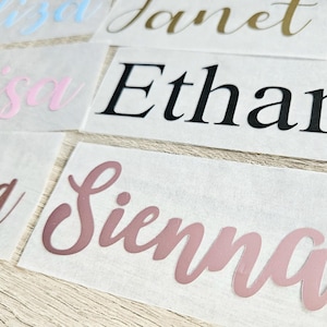 May include: Close-up of several name decals in various colors. Names include "Eliza" in light blue, "Janet" in gold, "Louisa" in pink, "Ethan" in black, "Amelia" in brown, and "Sienna" in mauve. Each name is in a cursive font.