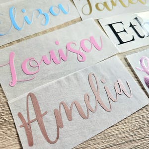 May include: A collection of name decals in various colors, including blue, pink, gold, black, and rose gold. Each name is displayed in a cursive font on a clear backing, perfect for personalizing items. The names include Eliza, Louisa, Amelia, and others.