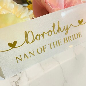 May include: A white card with gold lettering that reads "Dorothy Nan of the Bride" with two gold hearts on either side of the text.