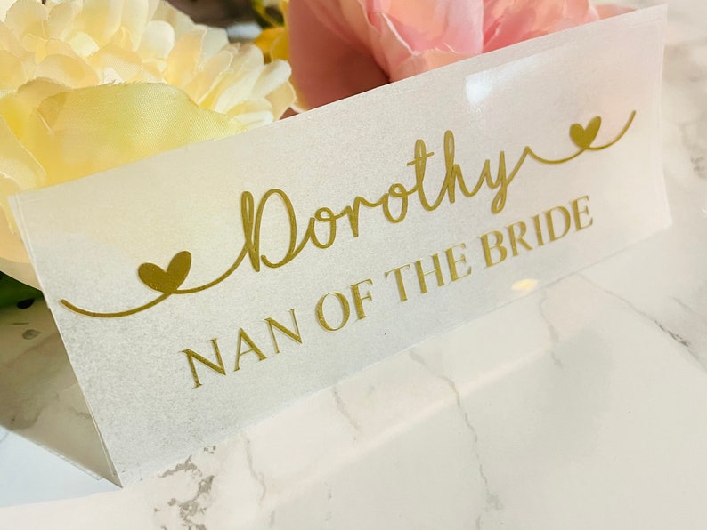 May include: A white card with gold lettering that reads "Dorothy Nan of the Bride" with two gold hearts on either side of the text.