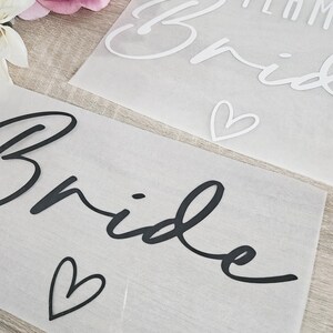 K&ouml;nnte beinhalten: Zwei transparente Bl&auml;tter mit dem Wort "Bride" in schwarzer Schrift und einem Herzsymbol. Ein Blatt hat die Worte "Team Bride" in Wei&szlig;. Hochzeitsthema.