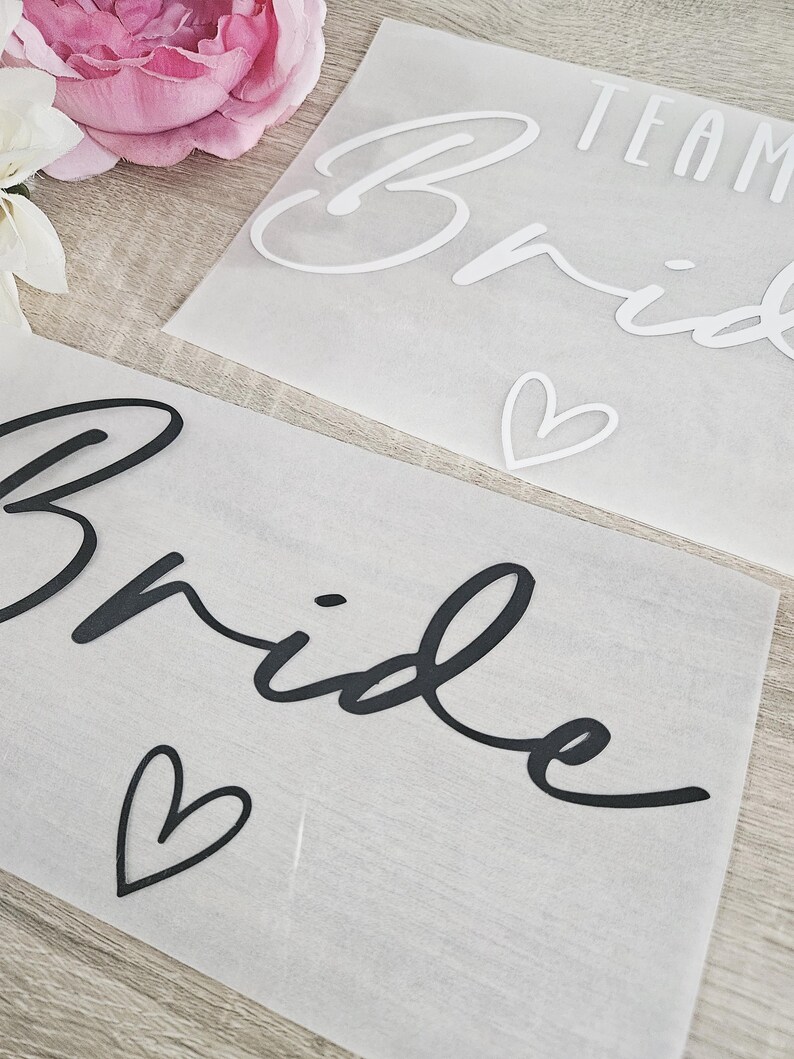K&ouml;nnte beinhalten: Zwei transparente Bl&auml;tter mit dem Wort "Bride" in schwarzer Schrift und einem Herzsymbol. Ein Blatt hat die Worte "Team Bride" in Wei&szlig;. Hochzeitsthema.