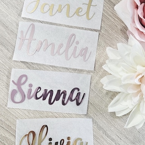 May include: Four name decals: "Janet" in gold, "Amelia" in pink, "Sienna" in purple, and "Olivia" in gold. Each name is in a cursive font, affixed to a white rectangular label. Decorative flowers are in the background.
