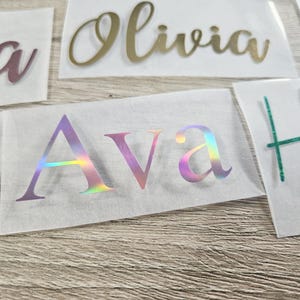 May include: Close-up of several name decals in various colors. The name "Ava" is in a holographic finish, while "Olivia" is gold. Other names are in pink, black, and teal. The decals are on a white backing.