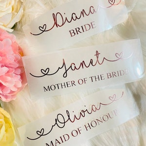 May include: Three white labels with rose gold lettering and hearts. The top label reads "Diana Bride". The middle label reads "Janet Mother of the Bride". The bottom label reads "Olivia Maid of Honour".