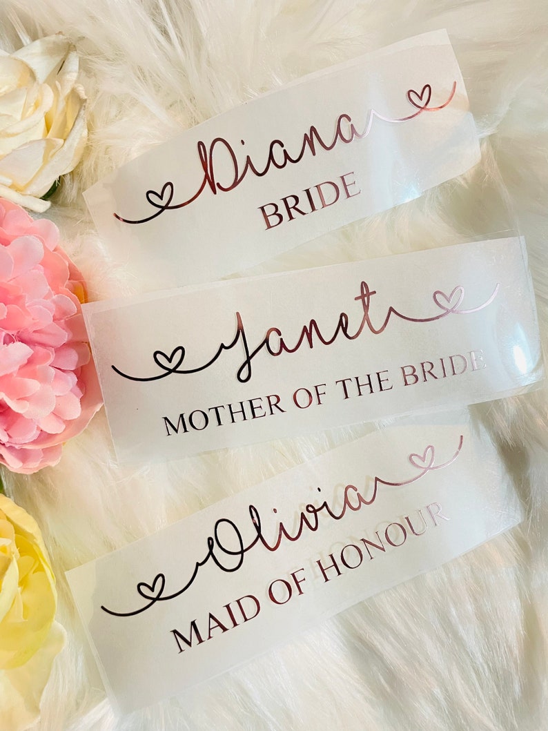 May include: Three white labels with rose gold lettering and hearts. The top label reads "Diana Bride". The middle label reads "Janet Mother of the Bride". The bottom label reads "Olivia Maid of Honour".