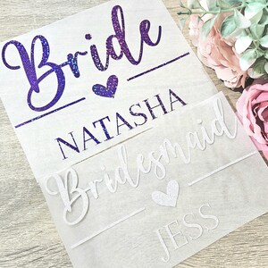 Op de afbeelding: Drie transparante vellen met tekst voor een bruiloft. Het bovenste vel leest "Bride" in paarse glitter met een hart en de naam "Natasha". Het onderste vel leest "Bridesmaid" en "Jess".