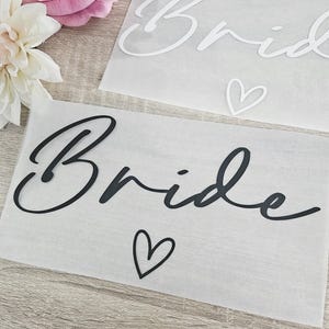 K&ouml;nnte beinhalten: Zwei Papierschnitte mit dem Wort "Bride" in Schreibschrift. Einer ist schwarz, der andere wei&szlig;. Unter dem schwarzen "Bride" befindet sich ein kleines Herz. Eine wei&szlig;e und rosa Blume befindet sich in der oberen linken Ecke.