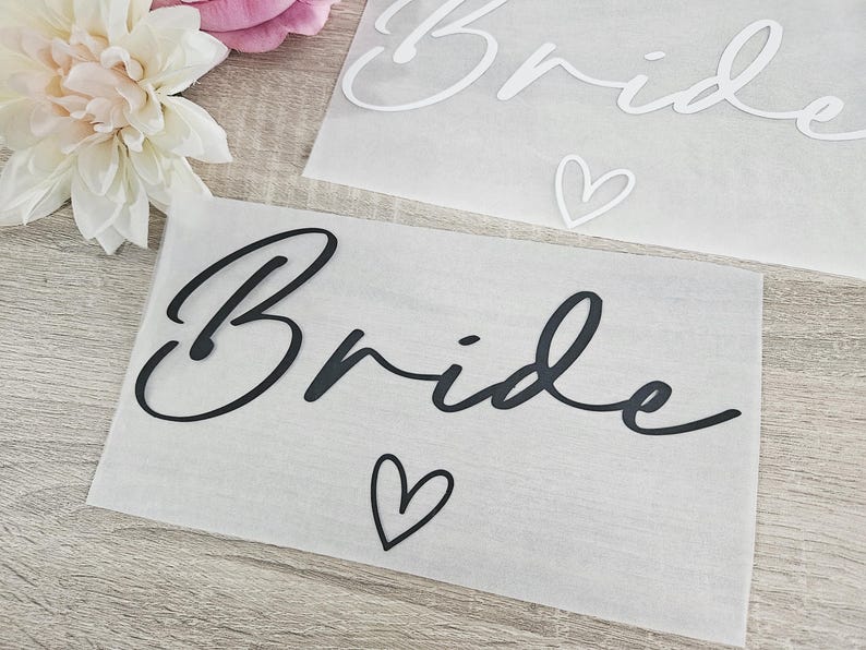 K&ouml;nnte beinhalten: Zwei Papierschnitte mit dem Wort "Bride" in Schreibschrift. Einer ist schwarz, der andere wei&szlig;. Unter dem schwarzen "Bride" befindet sich ein kleines Herz. Eine wei&szlig;e und rosa Blume befindet sich in der oberen linken Ecke.