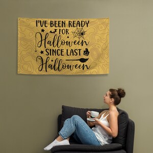 Puede incluir: Un tapiz de pared rectangular con temática de Halloween con el texto "I've Been Ready For Halloween Since Last Halloween" en negro. El fondo es un patrón tipo mapa en tonos amarillos y marrones. El diseño incluye un murciélago, una telaraña, una calabaza y una escoba.