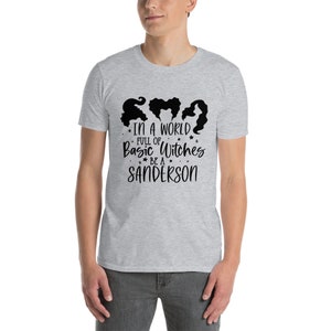 以下が含まれることがあります： 3 人の魔女のシルエットの黒いグラフィックと「IN A WORLD FULL OF Basic Witches BE A SANDERSON」というテキストが特徴のライトグレーの T シャツ。このシャツは柔らかい素材で作られています。