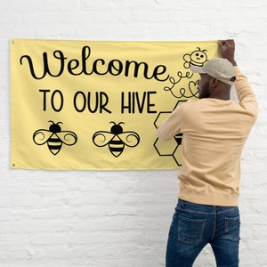 May include: A yellow banner with the text "Welcome to Our Hive" in black script. The banner features illustrations of bees and a honeycomb design. The banner is being held up against a white brick wall.