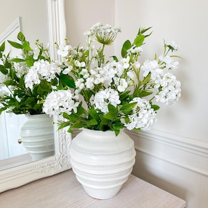 May include: A white ceramic vase with a ribbed design holds an arrangement of white flowers and green foliage. The vase sits on a light-coloured wooden surface, with a white framed mirror in the background.