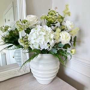 May include: A white ceramic vase with a ribbed design contains a display of white roses, hydrangeas, and greenery. The flowers are complemented by green foliage, creating a fresh and elegant arrangement. The vase is placed on a light-coloured wooden surface.