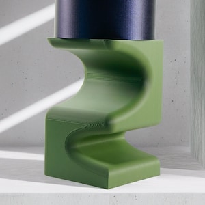 May include: A green, wavy plant stand with a dark blue planter and a leafy green plant with pink accents. The stand is on a white surface.