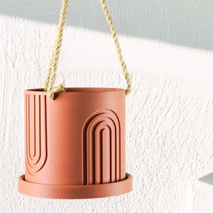 May include: A terracotta hanging planter with a geometric design. The planter is suspended from a natural rope.