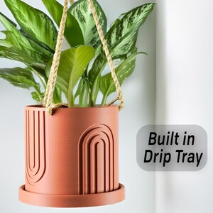 May include: A terracotta hanging planter with a built-in drip tray and a geometric design. The planter is hanging from a natural rope and has a green plant inside.