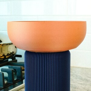 May include: A terracotta-colored bowl with a blue, pleated base. The bowl is sitting on a countertop.