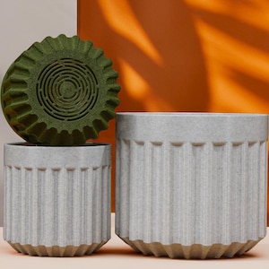 May include: Three decorative planters in a studio setting. Two are light gray with vertical ribbed detailing. The third is a dark green, circular object with a spiral design. The background is a mix of orange, gray, and peach.