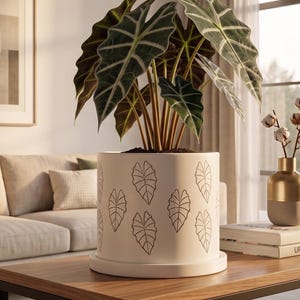 May include: A white ceramic plant pot with a leaf pattern contains a large plant with dark green leaves and light green veins. The pot sits on a wooden coffee table in a living room. A gold and beige vase with cotton stems is also on the table.
