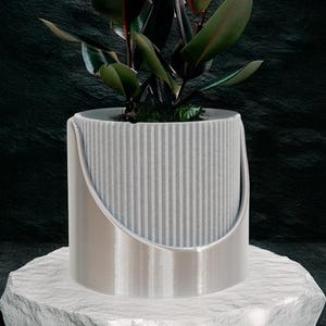 May include: A potted rubber plant with large, dark green leaves and reddish undersides. The plant is in a gray, ribbed planter with a silver metal handle, set on a white, textured stone. The background is a dark, textured surface.