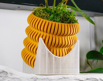 May include: A yellow, wavy, 3D printed planter with a white base holds a green plant with long, dark green leaves. The plant is in a white pot with moss around the base.
