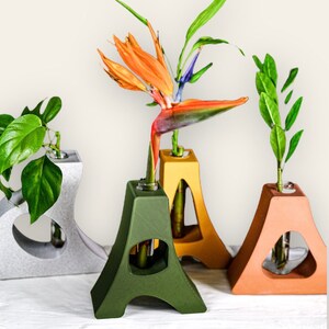 Puede incluir: Tres jarrones de cerámica en diferentes colores, gris, verde y naranja, con un diseño geométrico único. Cada jarrón tiene una pequeña abertura en la parte superior para flores. Una flor de ave del paraíso está en el jarrón verde, una planta de hojas está en el jarrón naranja y una planta de hojas está en el jarrón gris.