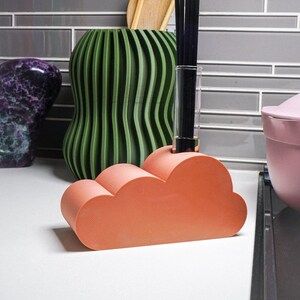 May include: A coral-colored cloud-shaped ceramic holder with a glass tube filled with black sticks. The holder is sitting on a countertop next to a green striped vase and a purple stone.