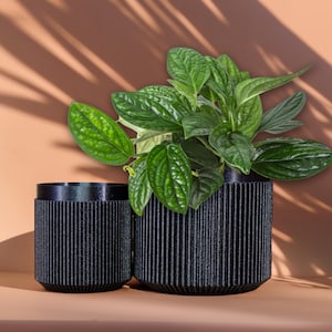 May include: Two black, ribbed plant pots of different sizes, one containing a lush green plant. The pots have a textured, vertical stripe design. The background is a warm, neutral colour with shadows.