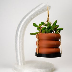 Può includere: Un portavasi in ceramica bianca con un braccio curvo che tiene un vaso sospeso in terracotta con una pianta succulenta.