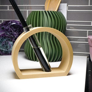 May include: A wooden stand with a circular opening holds two glass vials filled with dark liquid. The stand is light brown and has a smooth, rounded surface. The vials are clear and have a slightly textured surface. The stand is sitting on a white countertop.