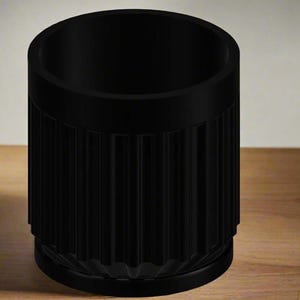 May include: A black cylindrical container with vertical ribbed detailing. The container has a flat base and a smooth rim. The object is made of a matte material and sits on a wooden surface. The overall design is minimalist and modern.