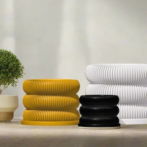 Peut inclure: Trois pots de fleurs ronds avec un design côtelé. Les pots sont jaunes, blancs et noirs. Les pots sont sur un sol en bois clair.