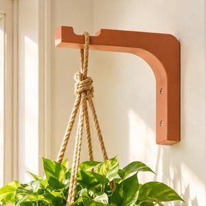 May include: A terracotta-colored L-shaped wall bracket mounted to a wall, supporting a hanging plant. A cream-colored ceramic pot holds a trailing pothos plant with variegated green and yellow leaves, suspended by a thick, natural-colored jute rope.