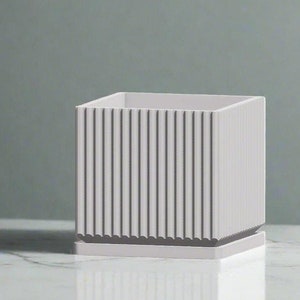 May include: A white square planter with a ribbed design. The planter has a small base and is made of a smooth material.
