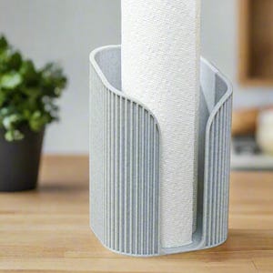 May include: A light gray paper towel holder with vertical ribbed detailing. A roll of white paper towels is placed inside the holder. The holder is on a wooden surface, with a potted plant and kitchen items in the background.