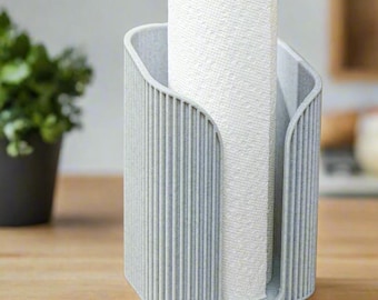 May include: A pale grey paper towel holder with vertical ribbed detailing. A roll of white paper towels is placed inside the holder. The holder is on a wooden surface, with a potted plant and kitchen items in the background.