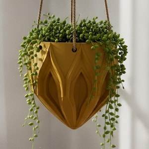 May include: A mustard yellow hanging planter with a geometric, faceted exterior design. The planter is suspended by three thin brown cords and contains a trailing string of pearls plant with small, round, green succulent leaves.