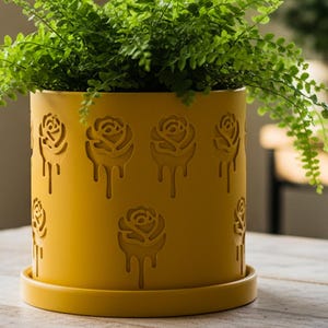 May include: A yellow ceramic planter with a fern. The planter has a cylindrical shape with a matching saucer. The planter is decorated with a rose design. The fern has bright green, delicate leaves. The planter is on a wooden table.