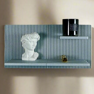 May include: A gray, textured wall shelf with two shelves. The top shelf has a black candle with a label that reads "Fragrance Noir - Aromatic Blue & Blackberry", and the bottom shelf has a white plaster bust of a man and a gold metal object.
