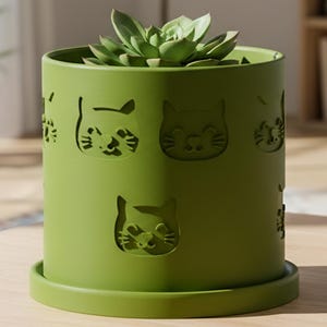 May include: A green cylindrical planter with cat face cutouts, holding a succulent plant. The planter sits on a matching saucer. The pot is on a light wood table, with other potted plants in the background.