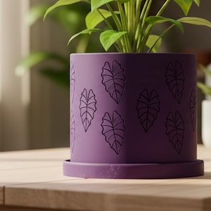 May include: A purple plant pot with a leaf pattern holds a green plant. The pot sits on a wooden table, with other small plants in the background. The pot has a matching saucer.