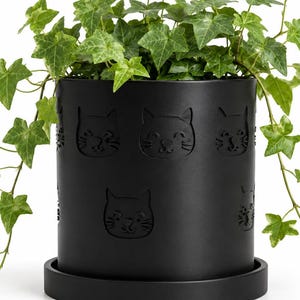 Könnte beinhalten: Ein schwarzer Blumentopf mit passender Untertasse, verziert mit geprägten Katzenköpfen. Der Topf enthält eine grüne Efeupflanze. Das Design ist modern und eignet sich für den Innen- und Außenbereich.