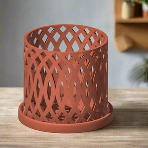 May include: A terracotta colored metal planter with a decorative lattice design. The planter has a round base and a cylindrical shape.