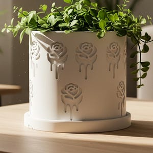 May include: A white cylindrical planter with a rose design, filled with green leafy plants, sits on a wooden table. Other potted plants are in the background. The planter has a matching saucer.